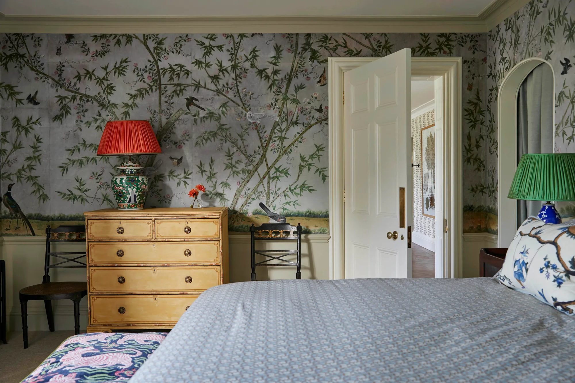 A guest bedroom with hand-painted Chinese wallpaper of birds and flowering branches, a painted chest of drawers and a coral lampshade