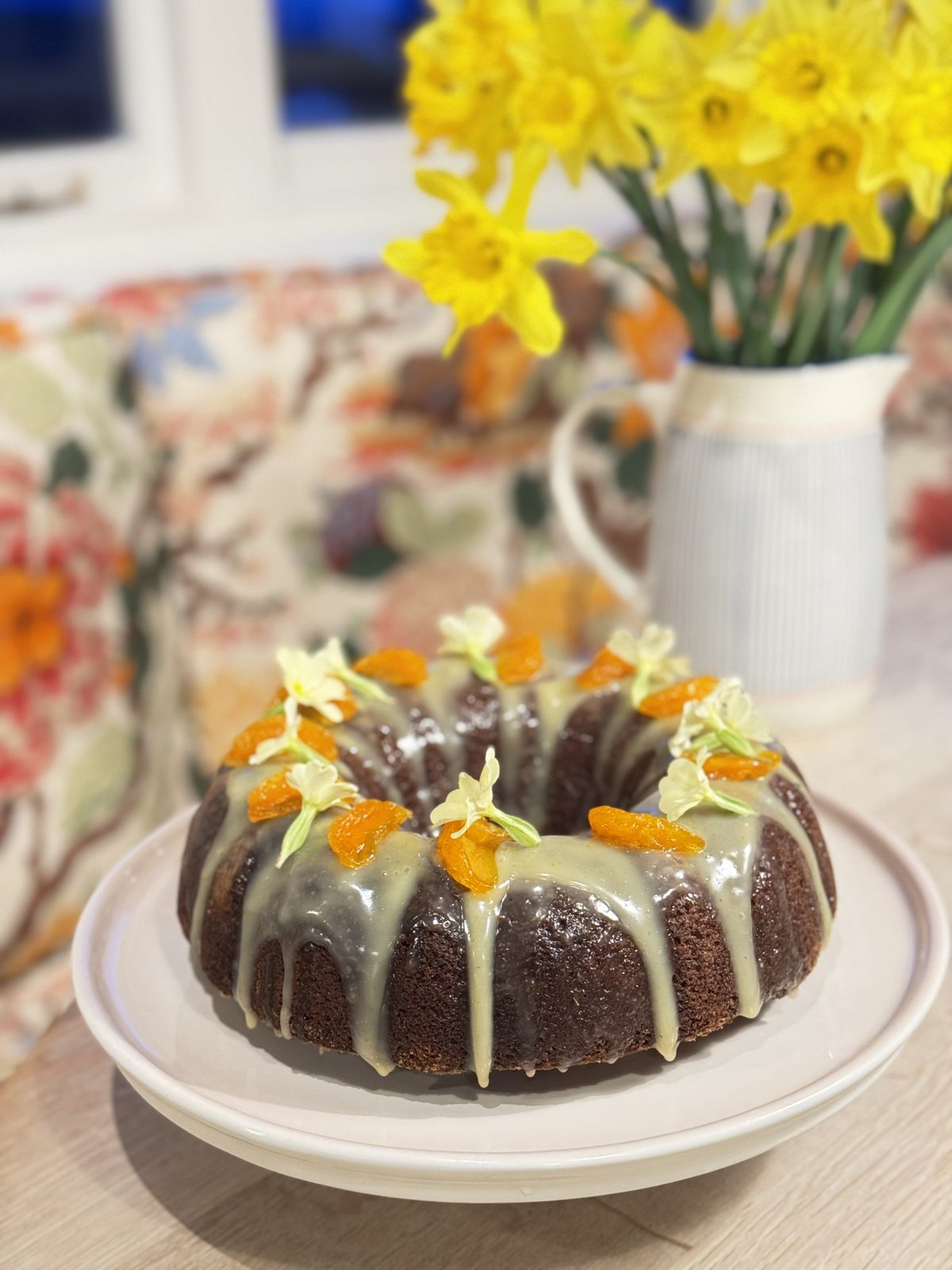 A ring cake with a pale glaze and candied orange, beside a jug of daffodils