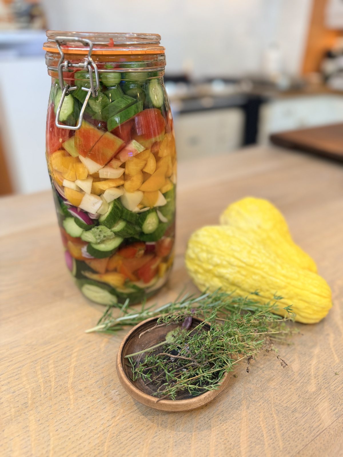 A tall jar of layered pickled vegetables with a patty-pan squash and a bowl of herbs